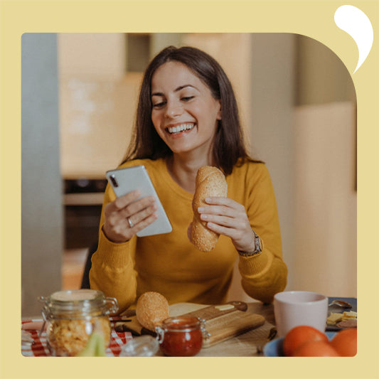 The young woman happily enjoys her breakfast, looking at her cell phone while holding her bread.